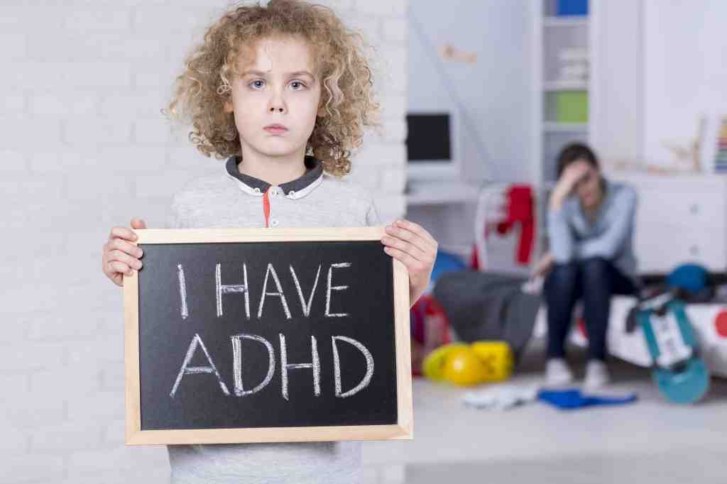 A kid holding a blackboard displaying "I have ADHD" while his mom is sad in the background.
